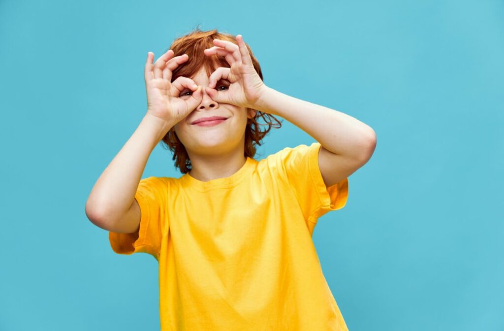 a child with a yellow shirt on a blue background is making his hands in the shape of glasses to symbolize eye care for children.
