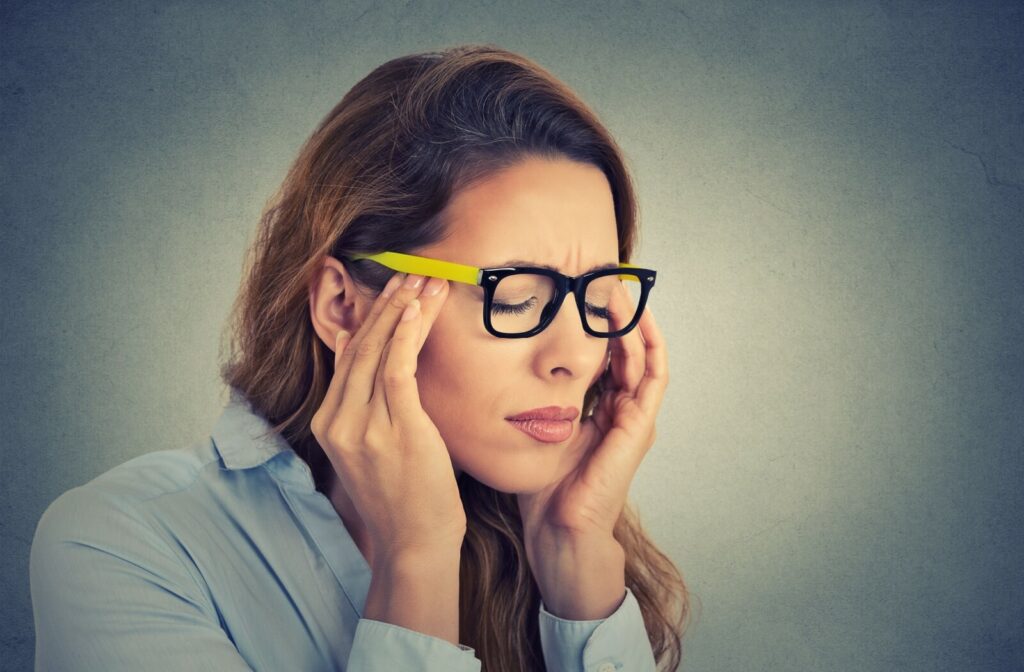 An adult wearing regular glasses rubs their temples as they feel a migraine coming on.