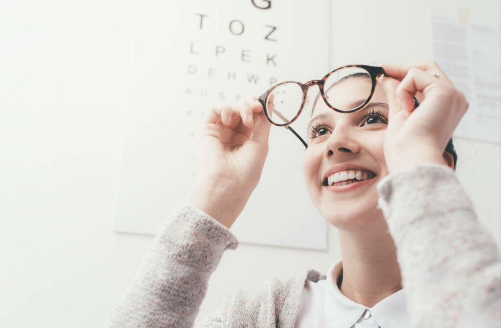 Smiling young person holding eyeglasses up to her face in an eye clinic, with a blurred Snellen eye chart visible in the background.