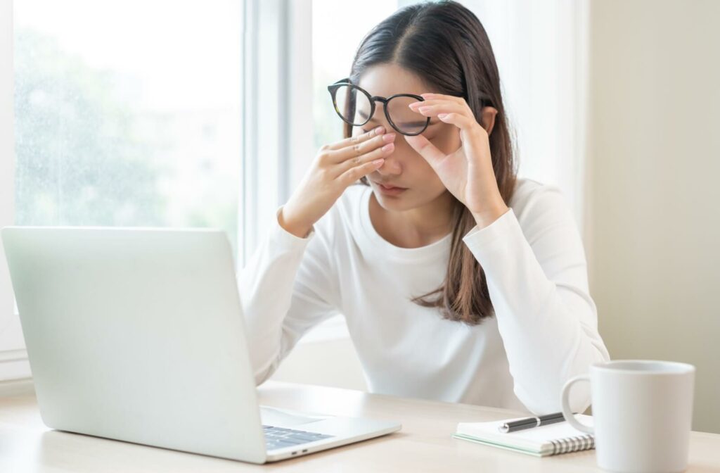 Person sitting at a desk with a laptop, rubbing her eyes in discomfort while holding her glasses, suggesting eye strain or dry eyes.
