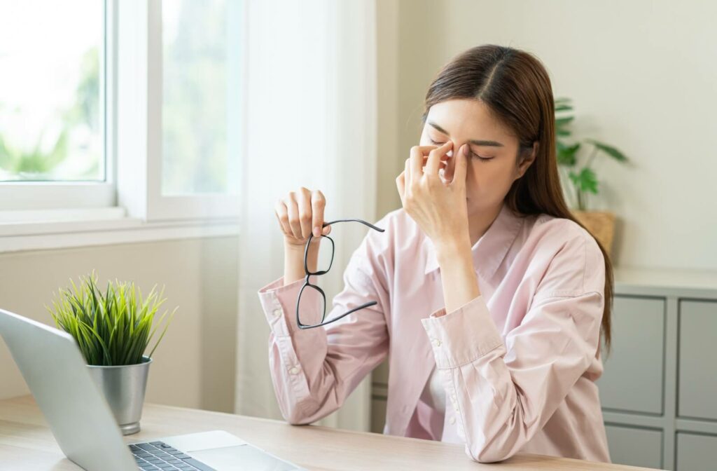 A person sitting at a desk with a laptop, eyes closed and rubbing the bridge of their nose while holding a pair of eyeglasses, demonstrating digital eye strain and symptoms of dry eye floaters.